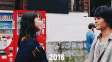 a man and a woman are standing in front of a red vending machine with the year 2016 on the bottom