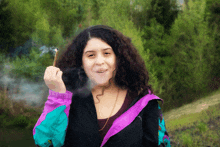 a woman with curly hair is smoking a cigarette in front of trees