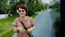 a woman wearing sunglasses and a flower in her hair smiles while walking down a path