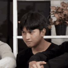 a young man in a black shirt is sitting in front of a window with a plant in a pot in the background .