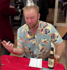 a man wearing a floral shirt sits at a table with a bottle and a glass of beer on it