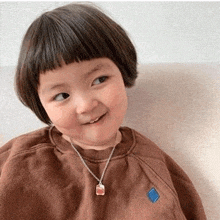 a little girl wearing a brown sweater and a necklace is sitting on a couch and smiling .