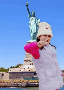 a little girl wearing a white hat with a star on it stands in front of the statue of liberty