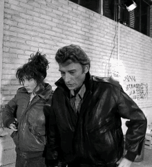 a man and a woman are standing in front of a brick wall with graffiti on it that says jvjv