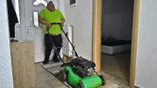 a man is pushing a green lawn mower down a hallway .