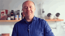 a man in a blue shirt is smiling with his eyes closed in front of a shelf with bottles and jars on it .