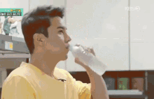 a man in a yellow shirt is drinking water from a bottle in a kitchen .