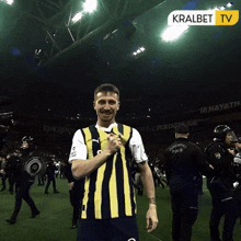 a man in a yellow and black striped shirt stands on a soccer field in front of a banner that says kralbet tv