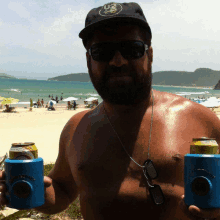 a shirtless man with a beard is holding two cans of beer on a beach