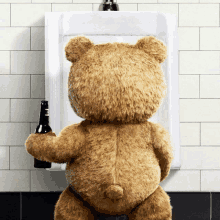 a teddy bear holding a bottle of beer standing in front of a urinal