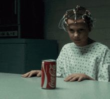 a child in a hospital gown sits at a table with a can of coca cola in front of her