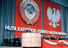 a man stands at a podium in front of a red curtain that says huta katowice-dzieko caleco narodu