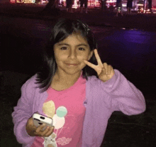 a little girl in a pink shirt is giving a peace sign while holding a cell phone .