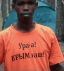 a man wearing an orange t-shirt with russian writing on it is standing in front of a blue tent .