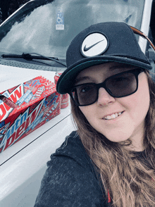 a woman wearing sunglasses and a hat is standing in front of a white truck with a box of mountain dew on the hood