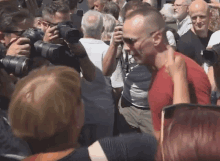 a man in a red shirt stands in a crowd of people taking pictures