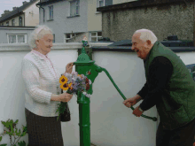 an elderly woman holds a bouquet of flowers while a man pumps water from a green pump