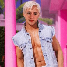 a shirtless man wearing a denim vest stands in front of a pink wall