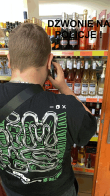 a man wearing a black shirt with a snake on it is talking on a cell phone in front of a shelf of alcohol