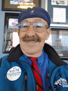 a man with glasses and a mustache wears a blue vest that says my vote before veteran on it