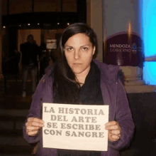 a woman in a purple jacket holds a sign that says la historia del arte se escribe con sangre