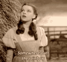 a black and white photo of a girl standing next to a pile of hay with the words `` somewhere over the rainbow '' .