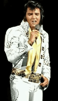 elvis presley is singing into a microphone while wearing a white suit and gold belt .