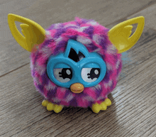 a pink and purple furby with yellow wings is sitting on a wooden floor