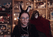 a woman wearing bunny ears and glasses is sitting in front of a bookshelf with a display of stuffed animals