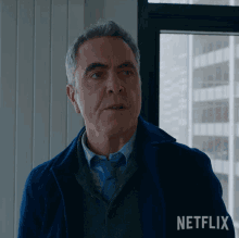 a man in a blue jacket stands in front of a window with a netflix logo behind him