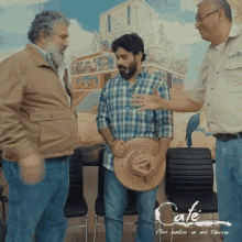 a man holding a straw hat talks to two other men in front of a sign that says cafe on it