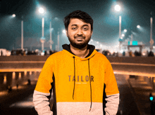 a man wearing a yellow tailor sweatshirt stands in front of a bridge