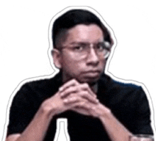 a man wearing glasses and a black shirt is sitting at a table with his hands folded in front of his face .