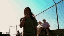 a woman is standing in front of a chain link fence while a group of people are sitting on a wall .