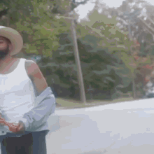 a man with a beard wearing a cowboy hat and a white tank top is walking down a street .