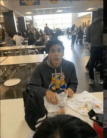 a young boy is sitting at a table in a cafeteria eating a sandwich .