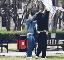 two people are flying a kite in a park with a trash can in the background that says ' recycle ' on it