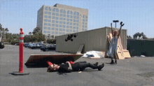 a man is laying on the ground in a parking lot while a woman stands behind him holding a hammer