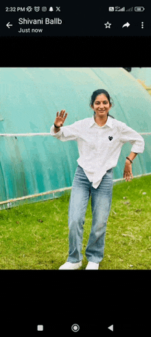 a woman in a white shirt and blue jeans is standing in front of a greenhouse and waving