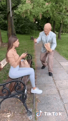 a woman is sitting on a bench talking to an older man who is holding a bag of food ..