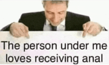 a man in a suit and tie is holding a white sign that says `` the person under me loves receiving anal '' .