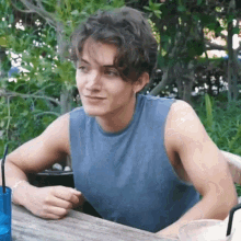 a young man in a blue tank top is sitting at a table with a drink in his hand .