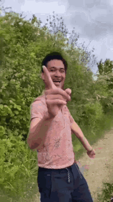 a man in a pink shirt is giving a peace sign while standing on a dirt road in the woods .