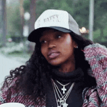 a woman wearing a hat and necklaces is sitting at a table with a cup of coffee in her hand .