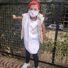 a little girl wearing a face mask and a white dress is standing in front of a fence .