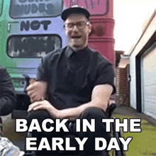 a man wearing glasses and a hat is sitting in front of a bus that says " back in the early day "