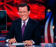 a man in a suit and tie is smiling while sitting at a desk in front of an american flag