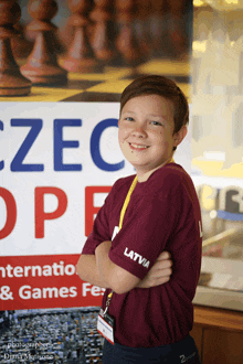 a boy wearing a latvia shirt stands in front of a chess board