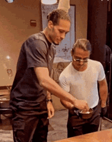 two men are standing next to each other at a table and one of them is pouring something into a bowl