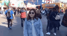 a woman wearing sunglasses and a denim jacket is standing in front of a crowd of people at an amusement park .